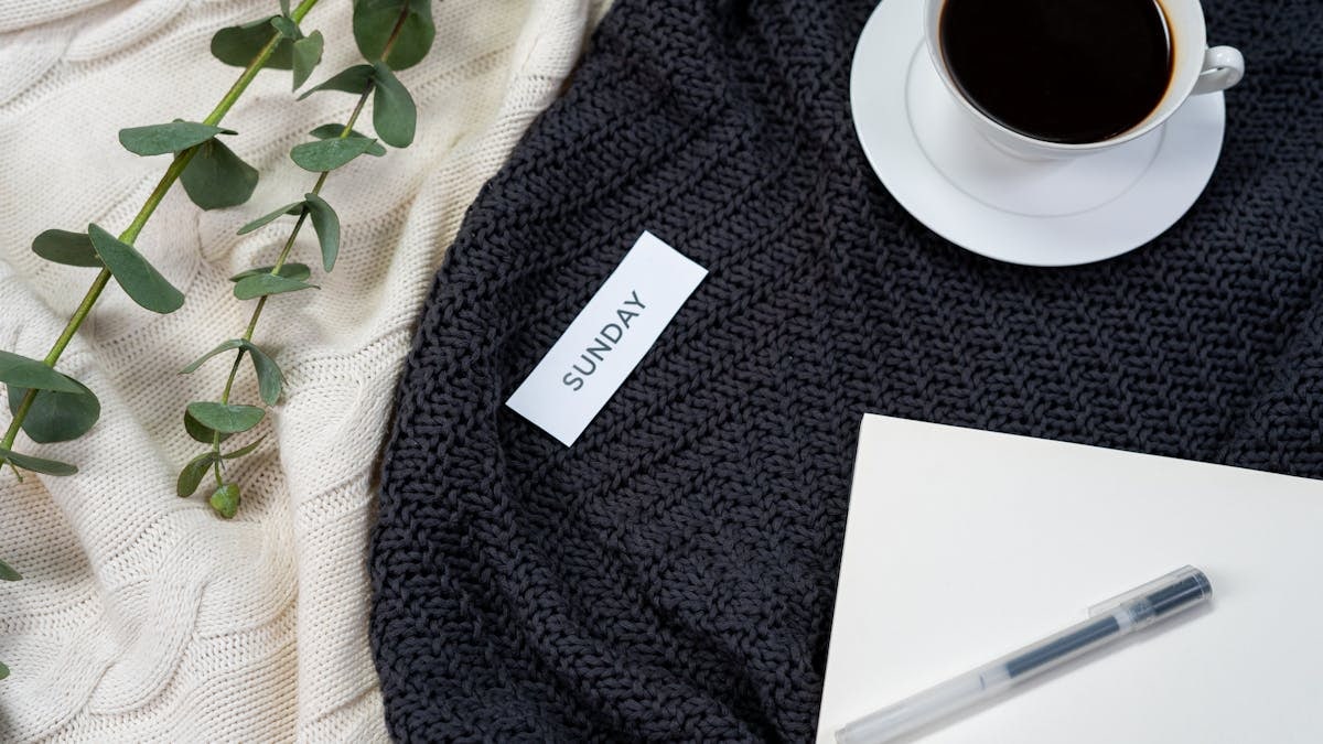 A flat lay of coffee, a knitted blanket, and a notepad creates a cozy Sunday atmosphere.